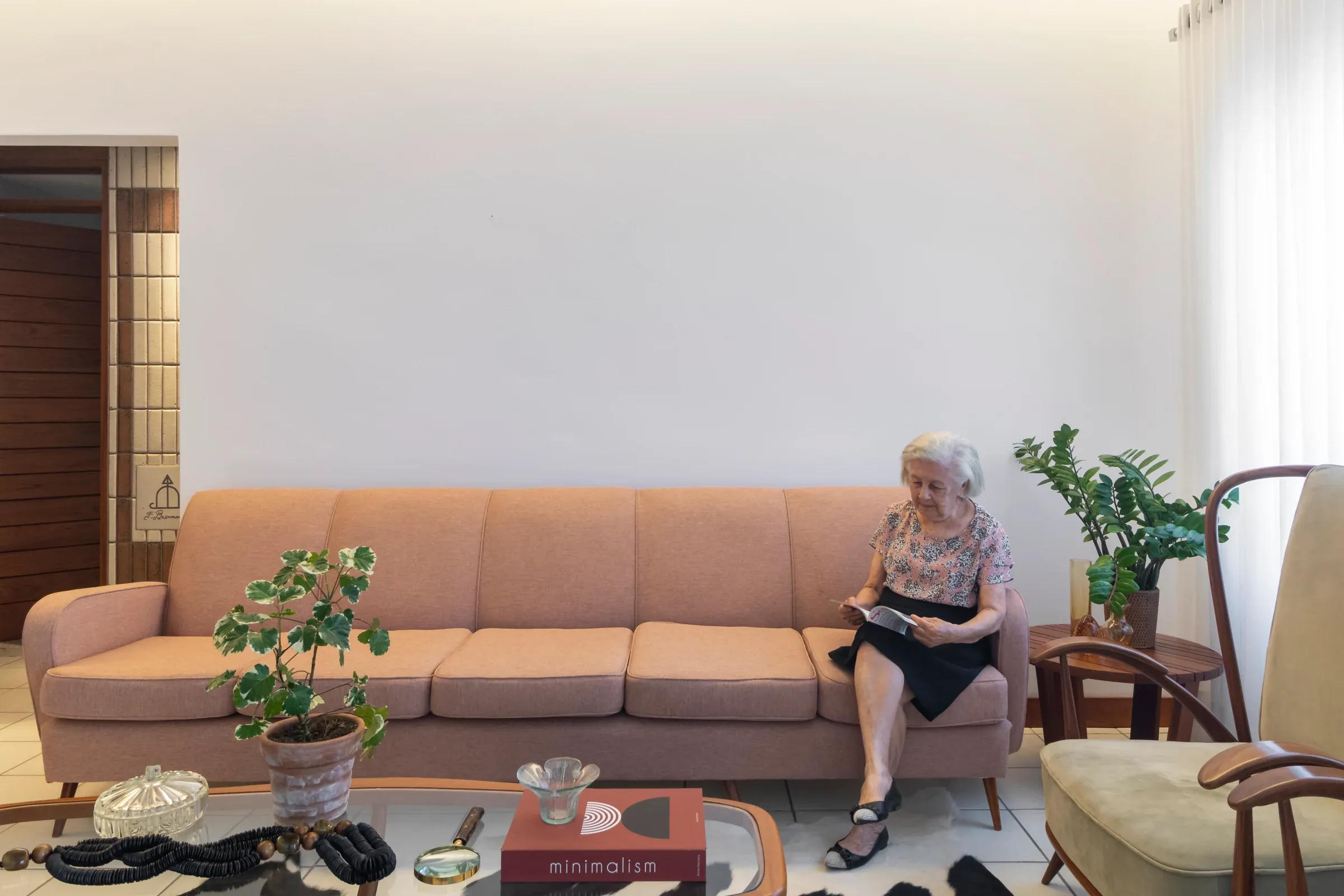Sala de estar acolhedora com moradora idosa lendo no sofá vintage rosa seco de quatro lugares, mesa de centro com livros e iluminação natural