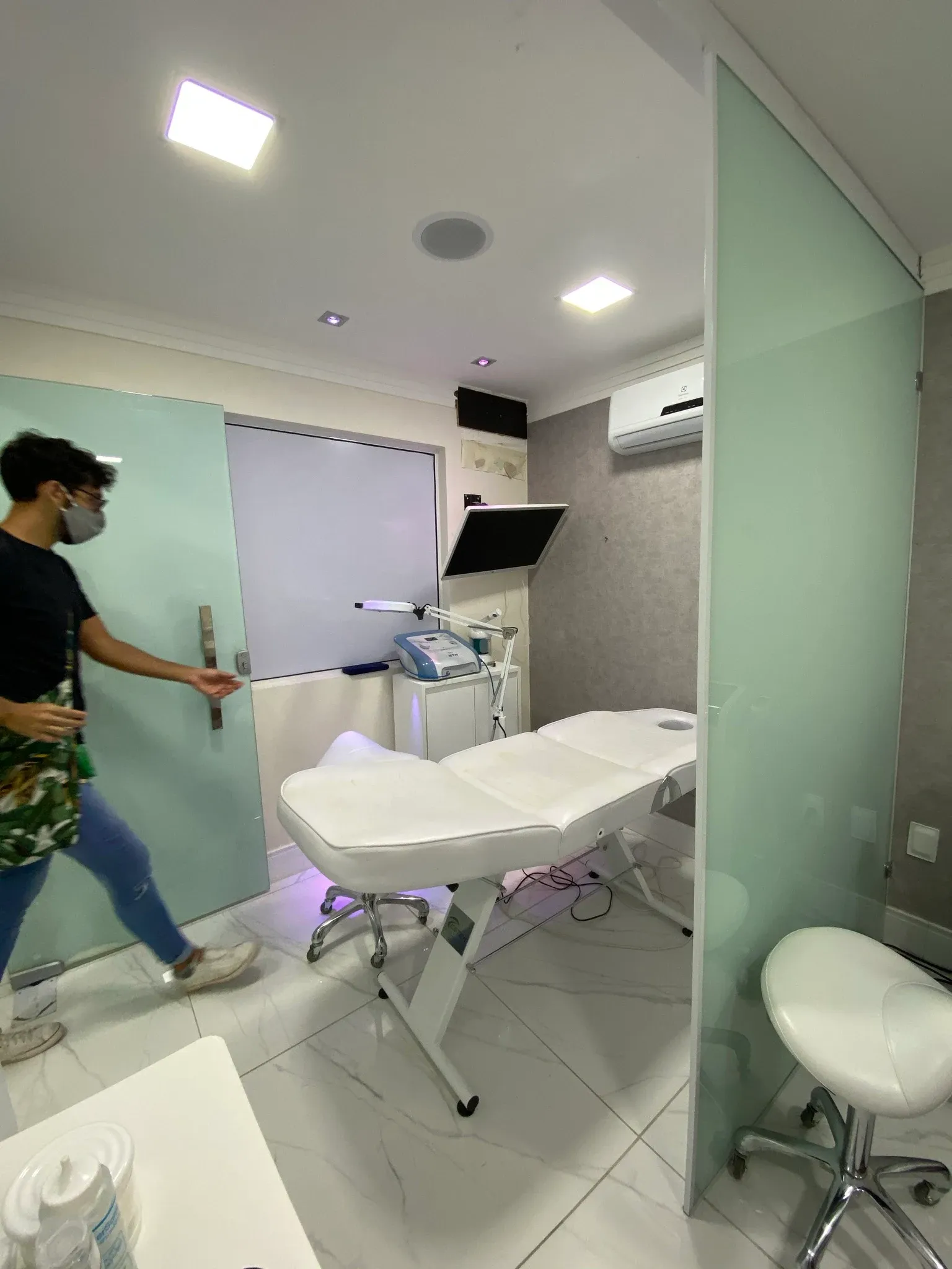 Cabine individual de procedimentos com divisórias em vidro fosco acidato, maca articulada reclinável, mocho ergonômico, equipamento de eletroterapia e ar-condicionado split